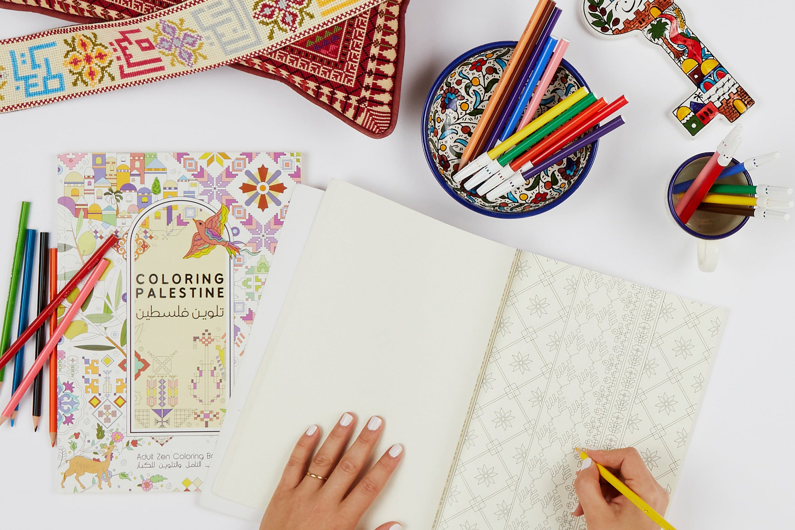 Person coloring in a book with a bowl of colored pencils and a coloring book titled 'Coloring Palestine' on a white surface.