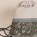 A drawstring backpack by ILHAAM. featuring a cream-colored top half with black branding, and a lower half with an intricate olive green, blue, and beige floral patterned fabric, set against a plain beige background.