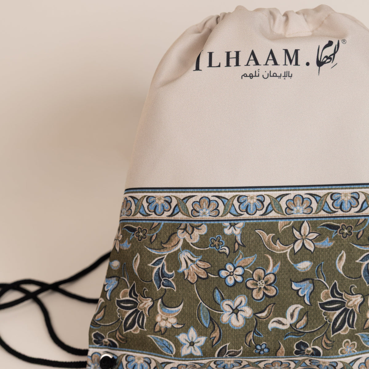 A drawstring backpack by ILHAAM. featuring a cream-colored top half with black branding, and a lower half with an intricate olive green, blue, and beige floral patterned fabric, set against a plain beige background.