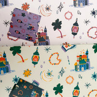 A split-screen view of two sheets of wrapping paper with a whimsical, colorful pattern of toy houses, lanterns, stars, and hearts on a white background. The left sheet has a lavender-purple patterned card placed on top, and the right sheet has a dark navy-blue patterned card placed on top.