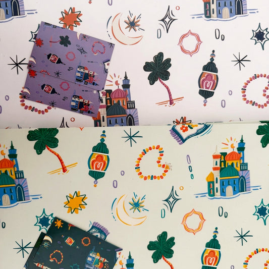 A split-screen view of two sheets of wrapping paper with a whimsical, colorful pattern of toy houses, lanterns, stars, and hearts on a white background. The left sheet has a lavender-purple patterned card placed on top, and the right sheet has a dark navy-blue patterned card placed on top.